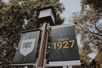 Two banners are displayed on a pole amidst tall, leafy trees. The banner on the left features a shield emblem with the year '1927' below it, while the one on the right prominently displays the text 'Since 1927' in bold letters.