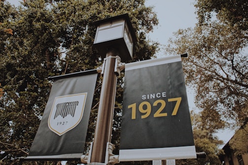 Two banners are displayed on a pole amidst tall, leafy trees. The banner on the left features a shield emblem with the year '1927' below it, while the one on the right prominently displays the text 'Since 1927' in bold letters.
