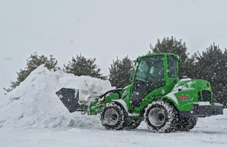 A green construction vehicle with large wheels is handling a pile of snow. The setting appears to be a snowy environment with falling snowflakes. Pine trees are visible in the background, adding a natural element to the wintry scene.
