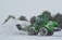 A green construction vehicle with large wheels is handling a pile of snow. The setting appears to be a snowy environment with falling snowflakes. Pine trees are visible in the background, adding a natural element to the wintry scene.