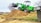A bright green compact loader sits atop a large mound of dirt, its scoop raised high in the air. The background features a few trees, some with sparse branches, and a row of commercial buildings, including a sign for a mattress center. A parked vehicle and a grassy field are also visible.