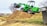 A bright green compact loader sits atop a large mound of dirt, its scoop raised high in the air. The background features a few trees, some with sparse branches, and a row of commercial buildings, including a sign for a mattress center. A parked vehicle and a grassy field are also visible.
