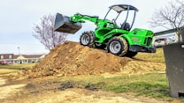 A bright green compact loader sits atop a large mound of dirt, its scoop raised high in the air. The background features a few trees, some with sparse branches, and a row of commercial buildings, including a sign for a mattress center. A parked vehicle and a grassy field are also visible.
