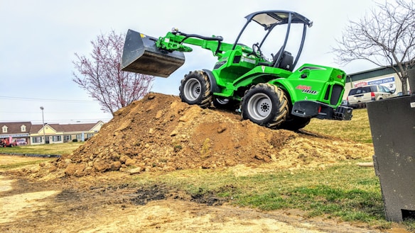 A bright green compact loader sits atop a large mound of dirt, its scoop raised high in the air. The background features a few trees, some with sparse branches, and a row of commercial buildings, including a sign for a mattress center. A parked vehicle and a grassy field are also visible.