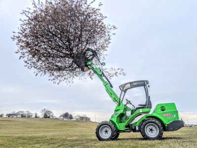 A bright green loader with a large scoop attachment is lifting a tree on a grassy field. The sky is overcast and the background features a few distant trees and small houses.