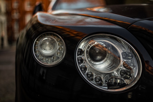 A close-up view of a car's headlight with intricate detailing and reflections. The headlight is round with multiple bulbs and a reflective surface, emphasizing its complexity and design.