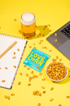 A bright, yellow background features a glass of beer with a frothy head, a small bowl filled with crunchy corn snacks, and a packet labeled 'LOVE CORN'. There's also an open laptop, a notebook with a pen resting on it, and scattered corn pieces throughout the scene.