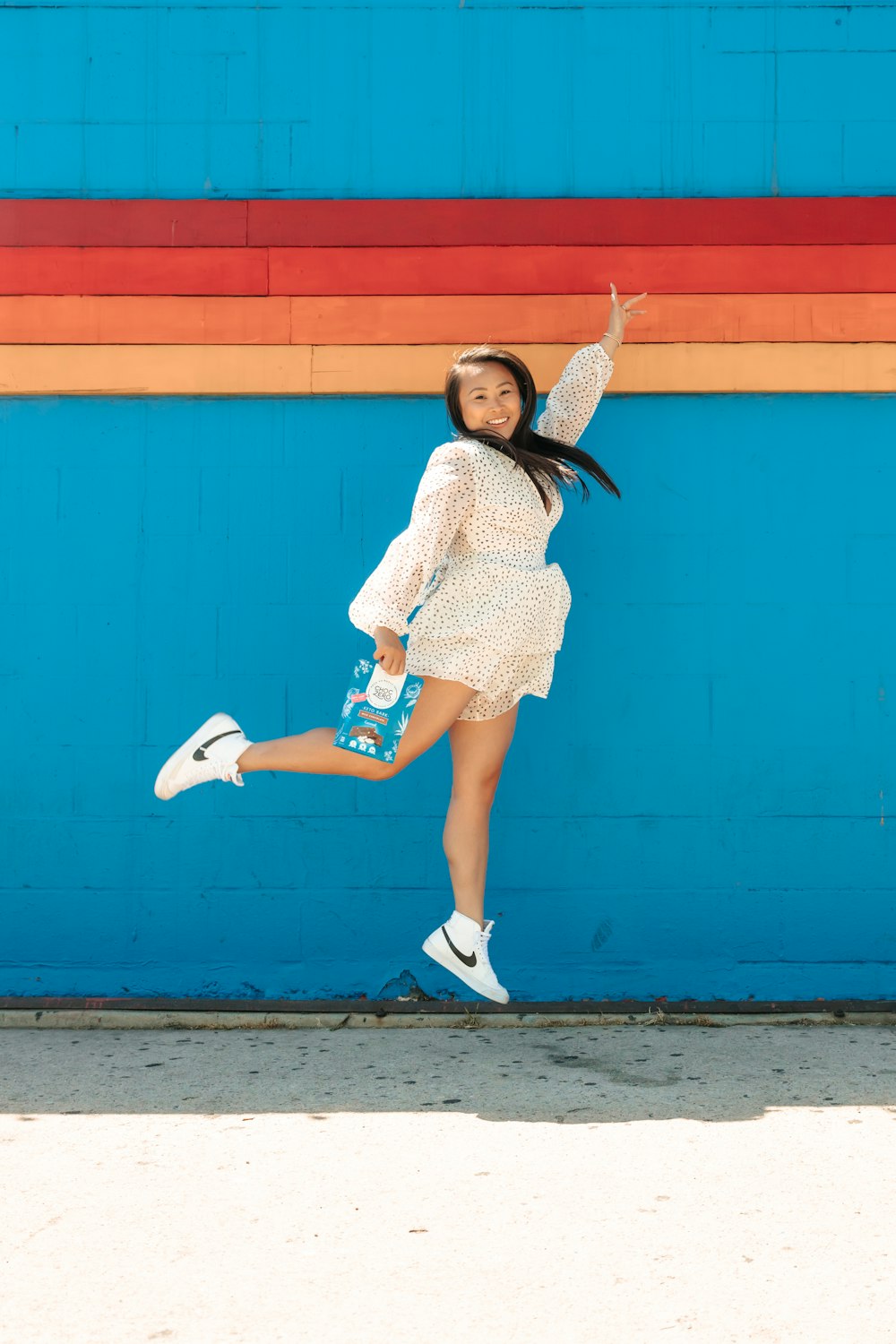 woman in white and black polka dot dress and white nike sneakers standing beside blue wall