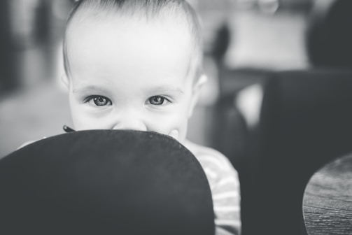 A baby peeks playfully over the edge of a seat or cushion, with only the upper part of the face visible. The close-up, black and white photograph captures the child's wide, curious eyes and soft skin, conveying a sense of innocence and curiosity.