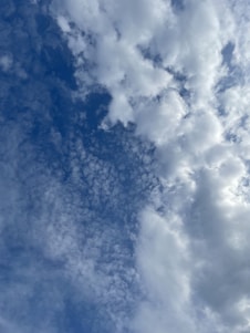 A cloudy sky featuring a variety of cloud formations, including fluffy cumulus clouds and smaller, scattered cirrus clouds all set against a backdrop of blue sky. The clouds range in density, creating a dynamic and textured appearance.