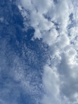 A cloudy sky featuring a variety of cloud formations, including fluffy cumulus clouds and smaller, scattered cirrus clouds all set against a backdrop of blue sky. The clouds range in density, creating a dynamic and textured appearance.