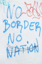 Graffiti on a wall with blue and red letters saying 'No Border No Nation'. There are additional scribbles and lines in black and red.