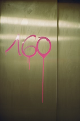 Pink graffiti numbers are painted on a metallic, possibly elevator, door with noticeable paint drips underneath the numbers.