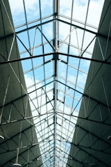A glass ceiling with metal beams and structures framing large, clear glass panels. The sky is visible through the ceiling, showing patches of blue with some clouds. The perspective is from below, looking up at the architectural structure.