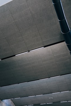 A modern ceiling with large rectangular panels and linear light fixtures. The panels are a uniform gray color, and the light fixtures create a symmetrical pattern. A large cylindrical vent or duct is visible in the upper corner, adding an industrial touch.