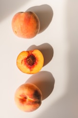 Three peaches are arranged on a light surface, casting soft shadows. Two whole peaches are positioned at the top and bottom, with their natural pinkish-orange hues. In the center, a halved peach reveals its vibrant orange flesh and textured pit.