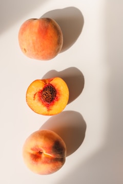 Three peaches are arranged on a light surface, casting soft shadows. Two whole peaches are positioned at the top and bottom, with their natural pinkish-orange hues. In the center, a halved peach reveals its vibrant orange flesh and textured pit.