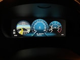 A close-up view of a car's digital dashboard display showing various information including a speedometer, a map indicating upcoming distance for navigation, a tachometer, temperature reading, and mileage. The speedometer is centered with a reading of 0 km/h, the tachometer is on the right showing the engine's revolutions per minute, and a digital map display with highlighted lanes and distance on the left. The dashboard display is illuminated in various shades of blue and white against a dark background.