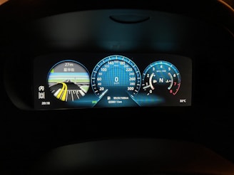A close-up view of a car's digital dashboard display showing various information including a speedometer, a map indicating upcoming distance for navigation, a tachometer, temperature reading, and mileage. The speedometer is centered with a reading of 0 km/h, the tachometer is on the right showing the engine's revolutions per minute, and a digital map display with highlighted lanes and distance on the left. The dashboard display is illuminated in various shades of blue and white against a dark background.