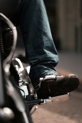 A close-up view of a person wearing dark leather shoes and denim jeans on a motorcycle footrest, capturing the detailed texture of the shoe and the shiny metallic parts of the bike.