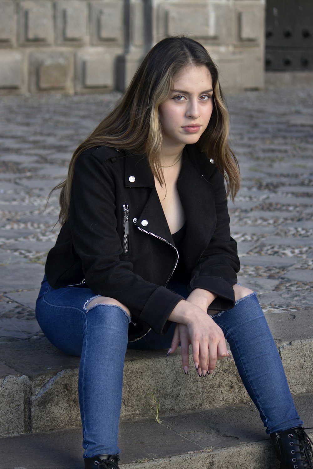 woman in black jacket and blue denim jeans sitting on gray concrete pavement