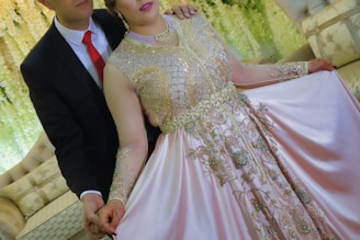 A couple is dressed in formal attire, with the woman wearing an ornate gown embellished with intricate embroidery and beading. The man is in a black suit with a red tie. They stand in a setting with floral decorations and elegant seating.