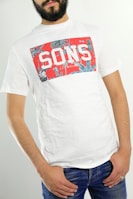 A person wearing a white t-shirt with a red, graphic design featuring the word 'SONS' in bold white letters. The t-shirt has a casual and relaxed fit, and the person pairs it with blue jeans. The individual is standing against a neutral background, with their hands in their pockets.