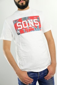 A person wearing a white t-shirt with a red, graphic design featuring the word 'SONS' in bold white letters. The t-shirt has a casual and relaxed fit, and the person pairs it with blue jeans. The individual is standing against a neutral background, with their hands in their pockets.