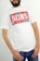 A person wearing a white t-shirt with a red, graphic design featuring the word 'SONS' in bold white letters. The t-shirt has a casual and relaxed fit, and the person pairs it with blue jeans. The individual is standing against a neutral background, with their hands in their pockets.