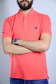 A person is standing with arms crossed wearing a coral polo shirt and a watch on their wrist. The logo on the shirt is blue and features a small bird.