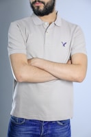 A person with a beard and wearing a light beige polo shirt stands with arms crossed. The shirt has a small emblem on the chest. The individual is also wearing blue jeans, and the background is a plain light blue.