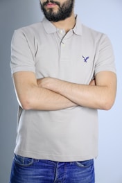 A person with a beard and wearing a light beige polo shirt stands with arms crossed. The shirt has a small emblem on the chest. The individual is also wearing blue jeans, and the background is a plain light blue.