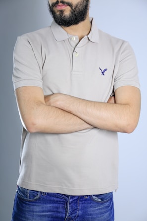 A person with a beard and wearing a light beige polo shirt stands with arms crossed. The shirt has a small emblem on the chest. The individual is also wearing blue jeans, and the background is a plain light blue.