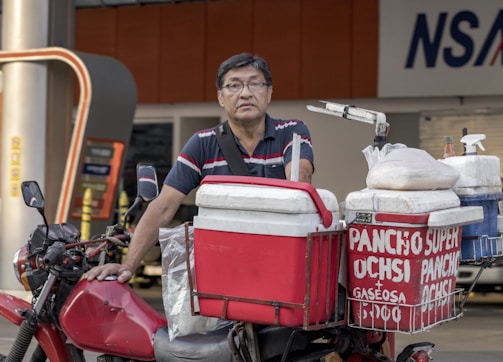 A man stands next to his motorcycle, which is equipped with several red and white coolers. The coolers are labeled with the words 'PANCHO SUPER' and 'GASEOSA' in white letters. The setting appears to be near a commercial area or street.