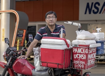 A man stands next to his motorcycle, which is equipped with several red and white coolers. The coolers are labeled with the words 'PANCHO SUPER' and 'GASEOSA' in white letters. The setting appears to be near a commercial area or street.