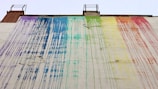A large wall features flowing streaks of paint in rainbow colors, with each hue blending into the next. The paint appears to have dripped from the roof, creating an abstract, artistic pattern. The building structure includes two ladder-like elements at the top.