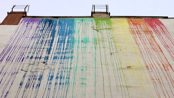 A large wall features flowing streaks of paint in rainbow colors, with each hue blending into the next. The paint appears to have dripped from the roof, creating an abstract, artistic pattern. The building structure includes two ladder-like elements at the top.
