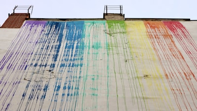A large wall features flowing streaks of paint in rainbow colors, with each hue blending into the next. The paint appears to have dripped from the roof, creating an abstract, artistic pattern. The building structure includes two ladder-like elements at the top.
