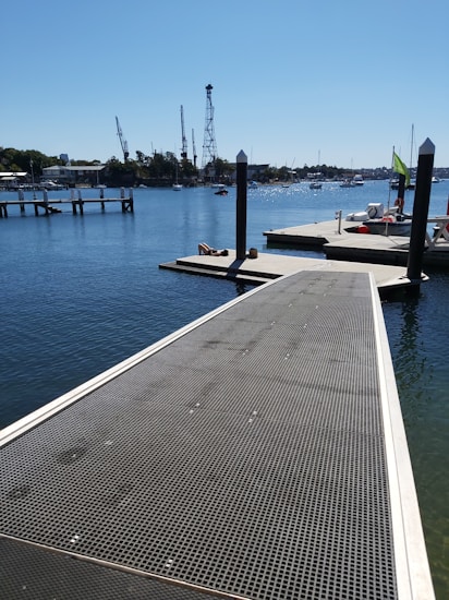 A metal dock leads into a bright, calm body of water, with boats moored to the sides and distant cranes visible against a clear blue sky. A person is sitting on the dock, and the area is surrounded by a serene harbor environment.