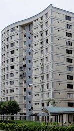 A tall residential building with multiple floors and a modern design, featuring light-colored tiles and blue-gray striped accents. The building has numerous windows and balconies, some with hanging laundry. A small structure with a transparent roof is visible at the base, surrounded by greenery, including trees and shrubs.