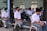 A group of students in school uniforms are seated in a classroom setting, wearing face masks. The setting appears to be an educational environment with students attentively waiting, some sitting on chairs and others on benches. The room has pale blue walls and windows with vertical blinds.