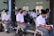 A group of students in school uniforms are seated in a classroom setting, wearing face masks. The setting appears to be an educational environment with students attentively waiting, some sitting on chairs and others on benches. The room has pale blue walls and windows with vertical blinds.