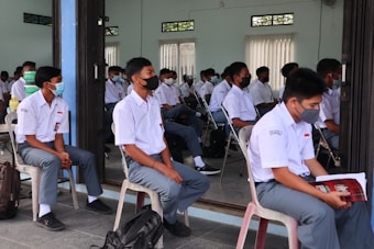 A group of students in school uniforms are seated in a classroom setting, wearing face masks. The setting appears to be an educational environment with students attentively waiting, some sitting on chairs and others on benches. The room has pale blue walls and windows with vertical blinds.