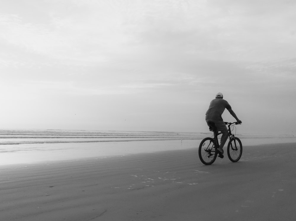grayscale photo of man riding bicycle on beach