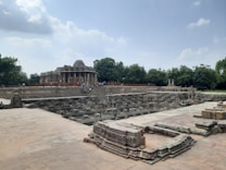 An ancient stepwell with intricate carvings is situated under a partly cloudy sky. In the background, a historic temple structure with detailed architecture is visible, surrounded by green trees. People are scattered throughout the site, examining the stonework.
