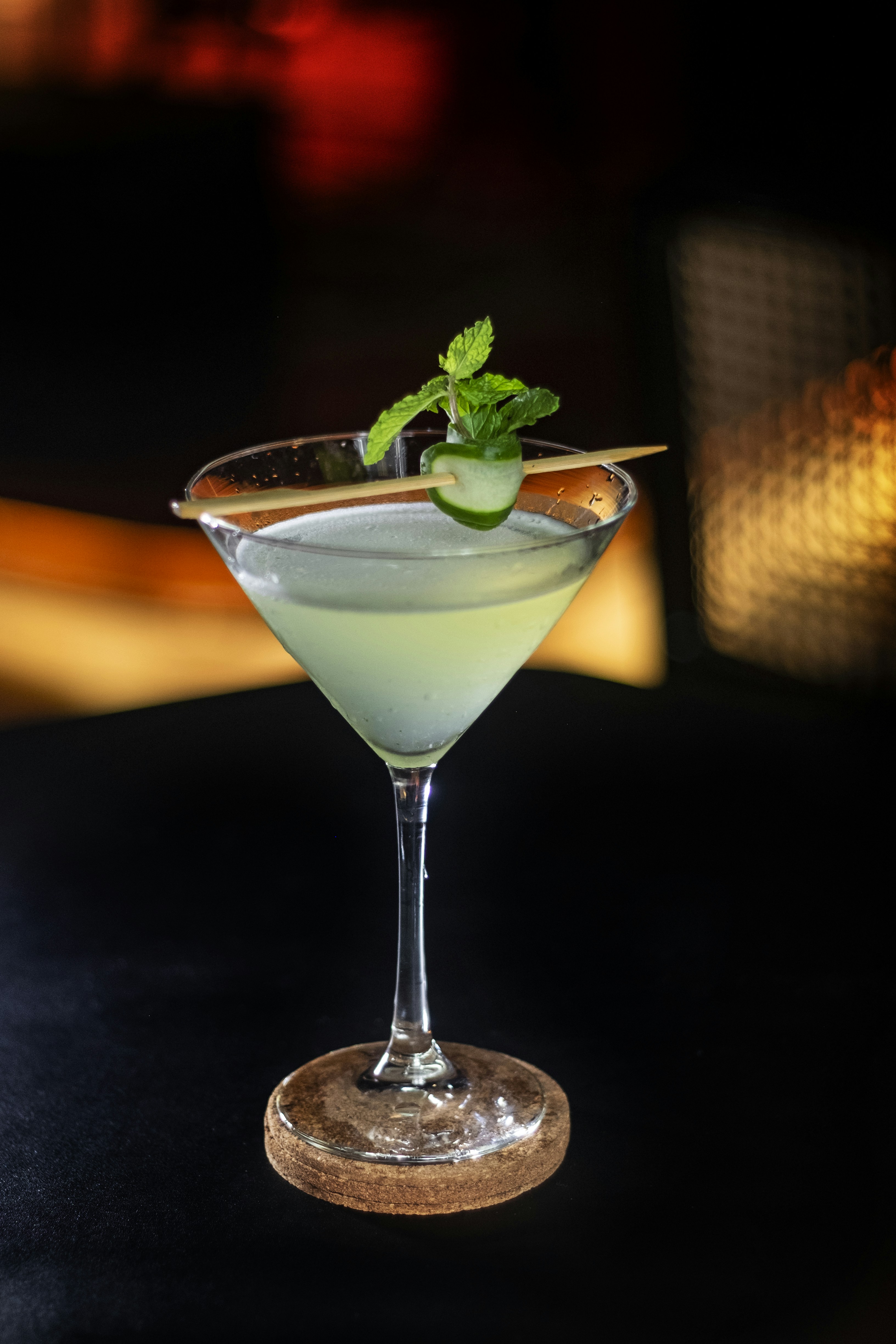 Green herbaceous cocktail with cucumber ribbon garnish, light mist, dark background