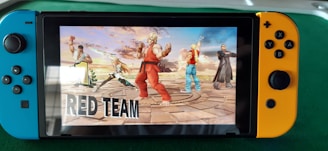 A handheld gaming console displays a screenshot of a fighting game featuring cartoonish characters in dynamic poses. The characters are standing in a virtual arena setting with a sky and cloud backdrop. The console is placed on a green surface.