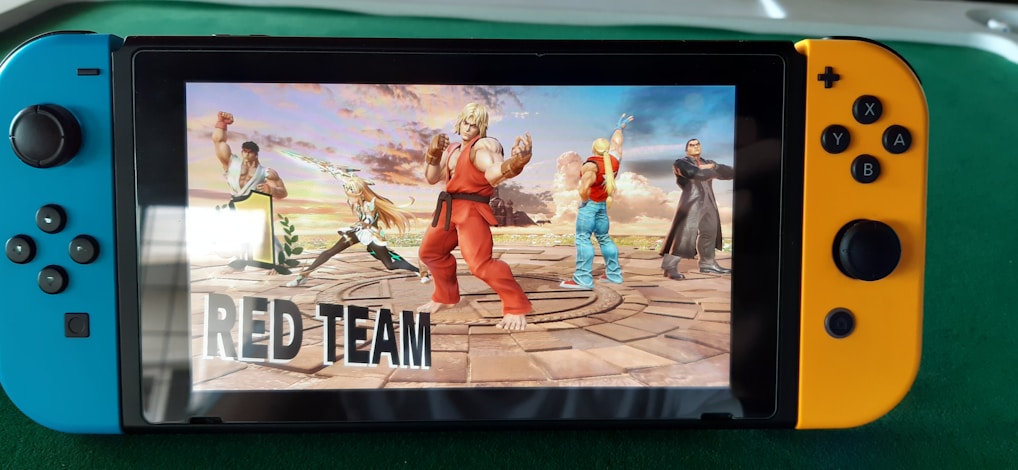 A handheld gaming console displays a screenshot of a fighting game featuring cartoonish characters in dynamic poses. The characters are standing in a virtual arena setting with a sky and cloud backdrop. The console is placed on a green surface.
