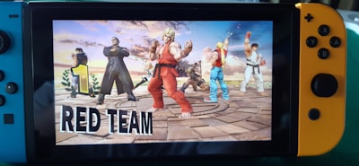 A Nintendo Switch console displays a fighting game with a group of characters standing victoriously on a circular platform. The main character in the foreground wears a red outfit, and the surrounding characters, each uniquely dressed, appear ready for combat. A large number 1 accompanied by the words 'RED TEAM' emphasizes their winning status.