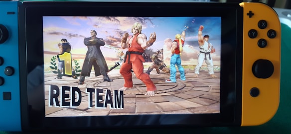 A Nintendo Switch console displays a fighting game with a group of characters standing victoriously on a circular platform. The main character in the foreground wears a red outfit, and the surrounding characters, each uniquely dressed, appear ready for combat. A large number 1 accompanied by the words 'RED TEAM' emphasizes their winning status.